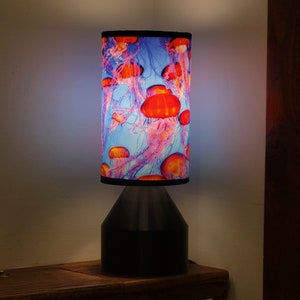 May include: A table lamp with a black base and a cylindrical lampshade featuring a colorful design of jellyfish in shades of pink, red, and blue.