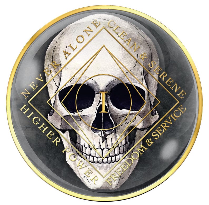 May include: A round, dark gray and gold emblem featuring a detailed skull illustration. The skull is centered within a gold geometric design, with text around the edges that reads "NEVER ALONE", "CLEAN & SERENE", "HIGHER POWER", and "FREEDOM & SERVICE".