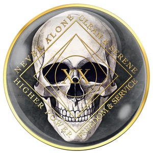 May include: A round, gray and gold medallion featuring a detailed skull illustration. The design includes gold geometric shapes and text that reads "NEVER ALONE", "CLEAN & SERENE", "HIGHER POWER", and "FREEDOM & SERVICE". The center displays "XX".