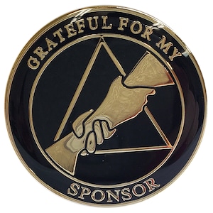 May include: A black and gold round pin with the text "Grateful for my Sponsor" and a gold triangle with a hand reaching out to another hand inside the triangle.