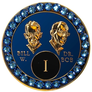 May include: A blue and gold round pin with a black circle in the center. The circle has a Roman numeral 'I' in white. The pin is surrounded by a ring of blue rhinestones. The words 'BILL W.' and 'DR. BOB' are printed on the pin.