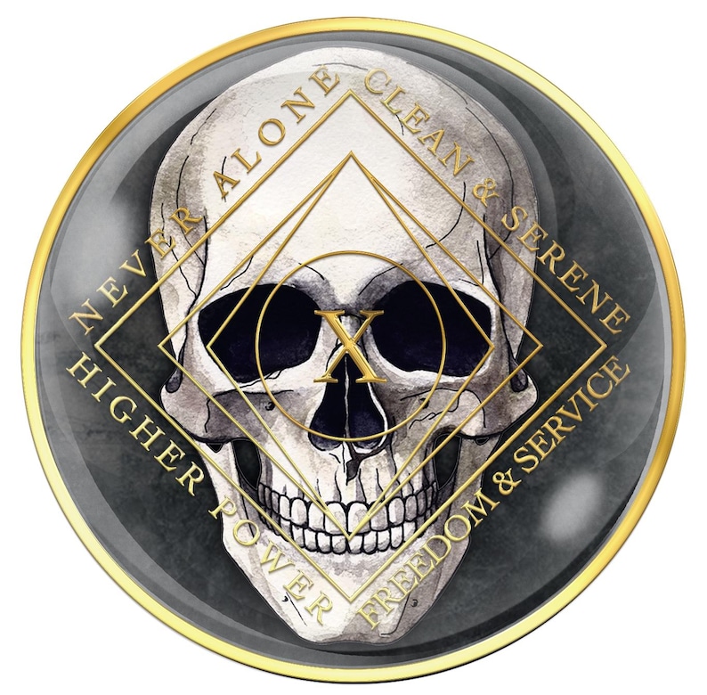 May include: A round, dark gray and gold-toned medallion featuring a detailed skull illustration. The skull is centered within a gold geometric design, with text around the edge that reads "NEVER ALONE, CLEAN & SERENE, HIGHER POWER, FREEDOM & SERVICE."