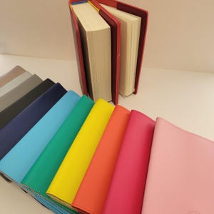 Puede incluir: Una colección de diarios y cuadernos coloridos. Las cubiertas son de varios colores, incluyendo rosa, rojo, naranja, amarillo, verde, azul, gris y negro. Un diario está abierto, revelando las páginas.