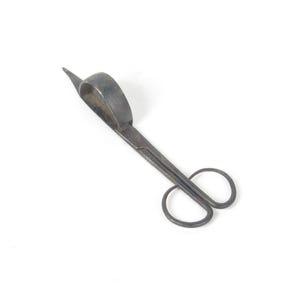 May include: Antique metal candle wick trimmer with a curved handle and finger loops. The scissor-like tool has a pointed blade and is designed to trim candle wicks. The metal has a dark, aged appearance.