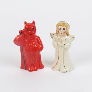 May include: Two ceramic figurines standing side by side. On the left, a bright red devil figure with horns and a black trident. On the right, a cream-colored angel figure with blonde hair, a halo, and wings, shown with hands clasped in a prayer position.