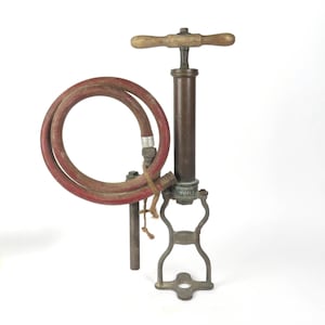 Puede incluir: Una bomba de bicicleta vintage de latón con un mango de madera y una manguera de goma roja. La bomba tiene una base con un marco de metal y una etiqueta que dice "HILL'S SPECIAL".