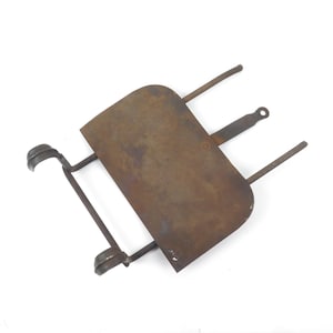 May include: A rectangular, weathered brown metal stand featuring a flat central plate, two curved supports on one side, and three extending metal rods on the opposite side, with a small circular hole at the end of the center rod.