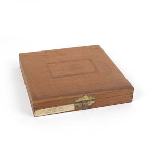 May include: A rectangular brown wooden storage box with a brass latch. The lid features a stamped label reading Kingsley Stamping Machine Co. Hollywood, California. A white paper label on the front edge reads H B M Goudy Cursive 18pt.