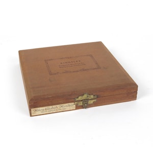 May include: A rectangular brown wooden storage box with a brass latch. The top features a printed label reading KINGSLEY Stamping Machine Co. Hollywood, California. A white paper label on the front reads Helene Elizabeth Hamilton GOUDY CURSIVE Lower Case.