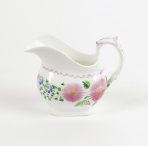 May include: A white porcelain creamer with a pink and green floral design. The creamer has a curved spout and a handle.