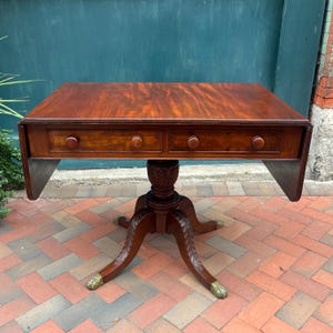May include: A rectangular mahogany drop-leaf table with a rich reddish-brown wood grain finish. The table features two drawers with round wooden knobs, a central pedestal base with carved leaf detailing, and four splayed legs with brass feet.