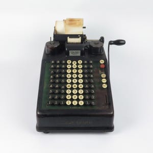 May include: A vintage black Burroughs Portable adding machine featuring a dark metal casing, a grid of numbered keys in cream and black, a side-mounted hand crank, and a paper roll holder at the top with a partially used roll of white paper.