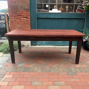 May include: A rectangular wooden table with a smooth, reddish-brown tabletop and a darker brown base. The table features four straight legs and two integrated drawers positioned horizontally along the front apron, each with a simple recessed handle.