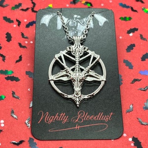 Peut inclure: Collier pendentif argent&eacute; avec un design de Baphomet &agrave; l'int&eacute;rieur d'un pentagramme. Le pendentif est sur une carte noire avec le texte "Nightly Bloodlust" en script rouge. Le fond est rouge avec des confettis en forme de chauve-souris.