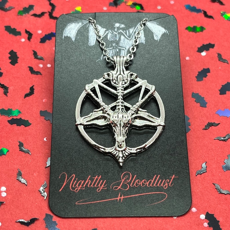Peut inclure: Collier pendentif argent&eacute; avec un design de Baphomet &agrave; l'int&eacute;rieur d'un pentagramme. Le pendentif est sur une carte noire avec le texte "Nightly Bloodlust" en script rouge. Le fond est rouge avec des confettis en forme de chauve-souris.