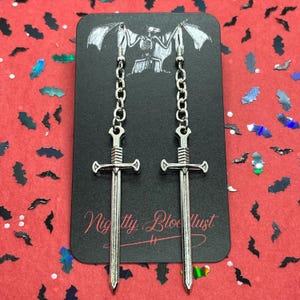 May include: Silver-colored sword earrings hanging from a chain. The earrings are displayed on a black card with a skeleton and bat wings illustration. The card has the text "Nightly Bloodlust" in cursive. The background is red with bat-shaped confetti.