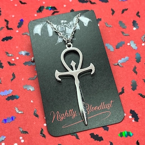 May include: A silver-colored ankh pendant necklace. The ankh is on a black card with the text "Nightly Bloodlust" in a cursive font. The background is red with bat-shaped confetti.
