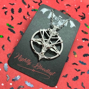 Peut inclure: Collier pendentif argent&eacute; avec un design de pentagramme et une t&ecirc;te de ch&egrave;vre au centre. Le pendentif est sur une carte noire avec le texte "Nightly Bloodlust" en script rouge. Le fond est rouge avec des confettis en forme de chauve-souris.