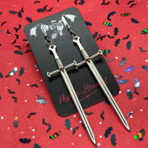 May include: A pair of silver sword earrings. The swords are crossed and have a detailed design. The earrings are hanging from a black card with the text "Night Blood Art" written on it.