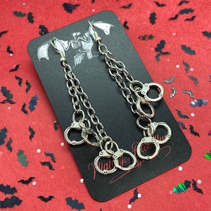 May include: Silver dangle earrings with a chain and multiple handcuff charms. The earrings are displayed on a black card with a bat design and the text "Night Culture".