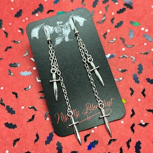 May include: Silver dangle earrings with a chain and a silver dagger charm. The earrings are displayed on a black card with the text "Nightly Bloodlust".