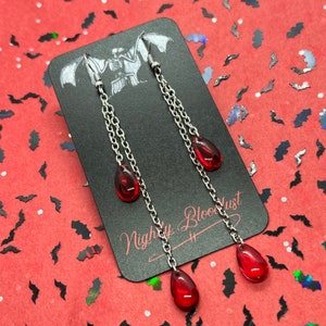 May include: A pair of silver chain earrings with three red teardrop-shaped charms. The earrings are displayed on a black card with the text "Nightly Bloodlust".