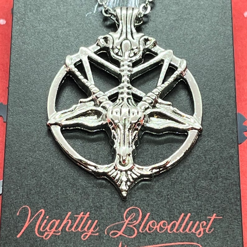 Peut inclure: Collier pendentif argent&eacute; avec un design de pentagramme d&eacute;taill&eacute; et une t&ecirc;te de ch&egrave;vre au centre. Le collier est suspendu &agrave; une cha&icirc;ne d&eacute;licate. Le texte "Nightly Bloodlust" est visible sur le fond noir.