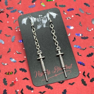 May include: A pair of silver syringe earrings with chain drop. The earrings are on a black card with the text "Nightly Bloodlust".