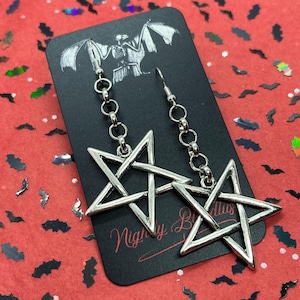 May include: Silver-toned pentagram earrings hanging from a chain. The earrings are displayed on a black card with a bat illustration and the text "Nighty By Nillus" on a red background with bat-shaped confetti.