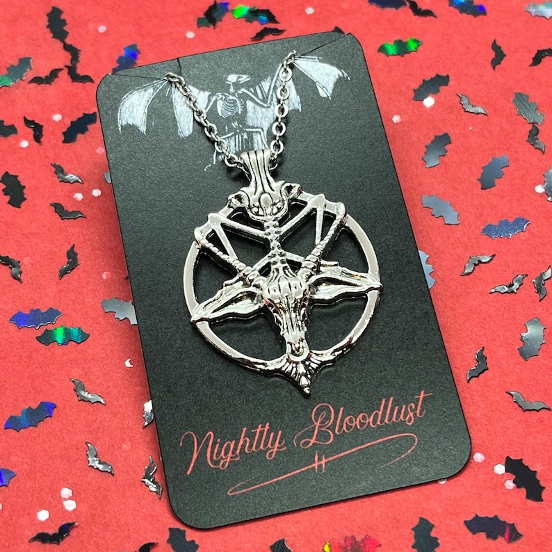 Peut inclure: Un collier pendentif argent&eacute; avec un design de Baphomet &agrave; l'int&eacute;rieur d'un pentagramme. Le collier est pr&eacute;sent&eacute; sur une carte noire avec le texte "Nightly Bloodlust". Le fond est rouge avec des confettis en forme de chauve-souris.