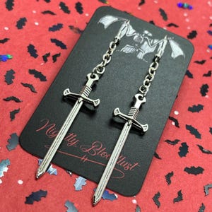 May include: Silver-colored sword earrings with chain detail. The earrings are displayed on a black card with the text "Nighty Bloodlust" and a bat-themed design. The background is red with bat-shaped confetti.