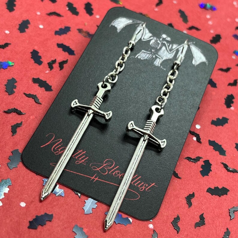 May include: Silver-colored sword earrings with chain detail. The earrings are displayed on a black card with the text "Nighty Bloodlust" and a bat-themed design. The background is red with bat-shaped confetti.