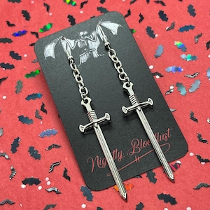 May include: Silver-colored sword earrings hanging from a chain, displayed on a black card with the text "Nightly Bloodlust". The card has a bat-like illustration at the top. The background is red with bat-shaped confetti.