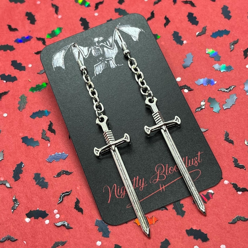 May include: Silver-colored sword earrings hanging from a chain, displayed on a black card with the text "Nightly Bloodlust". The card has a bat-like illustration at the top. The background is red with bat-shaped confetti.