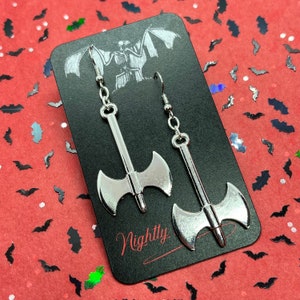 May include: A pair of silver axe earrings on a black card with the word "Nightty" printed on it. The earrings are hanging from silver hooks.