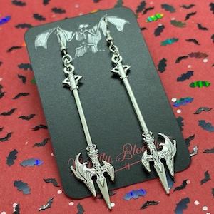 May include: A pair of silver-toned dangle earrings shaped like long, pointed spears or tridents with ornate, wing-like detailing at the base. The earrings are attached to a black display card featuring a white skeletal illustration and text.