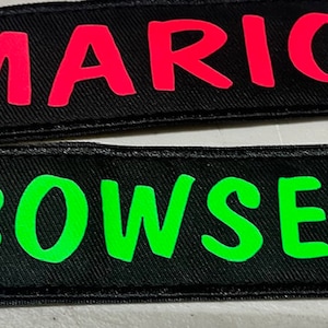 May include: Two black fabric patches with white stitching. The top patch has the word "MARIO" in bright pink letters. The bottom patch has the word "BOWSER" in bright green letters.