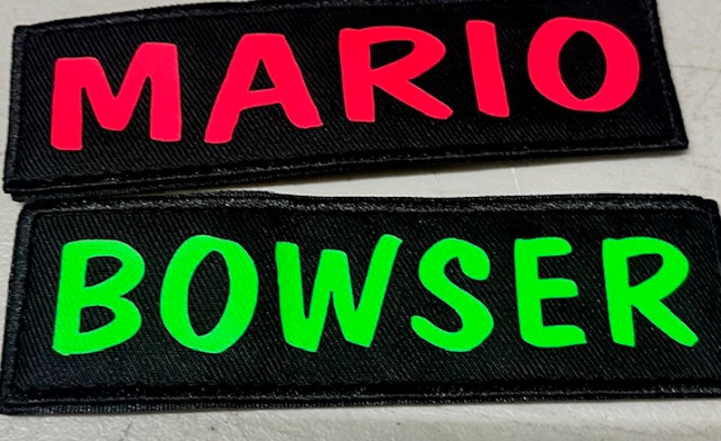 May include: Two black fabric patches with white stitching. The top patch has the word "MARIO" in bright pink letters. The bottom patch has the word "BOWSER" in bright green letters.