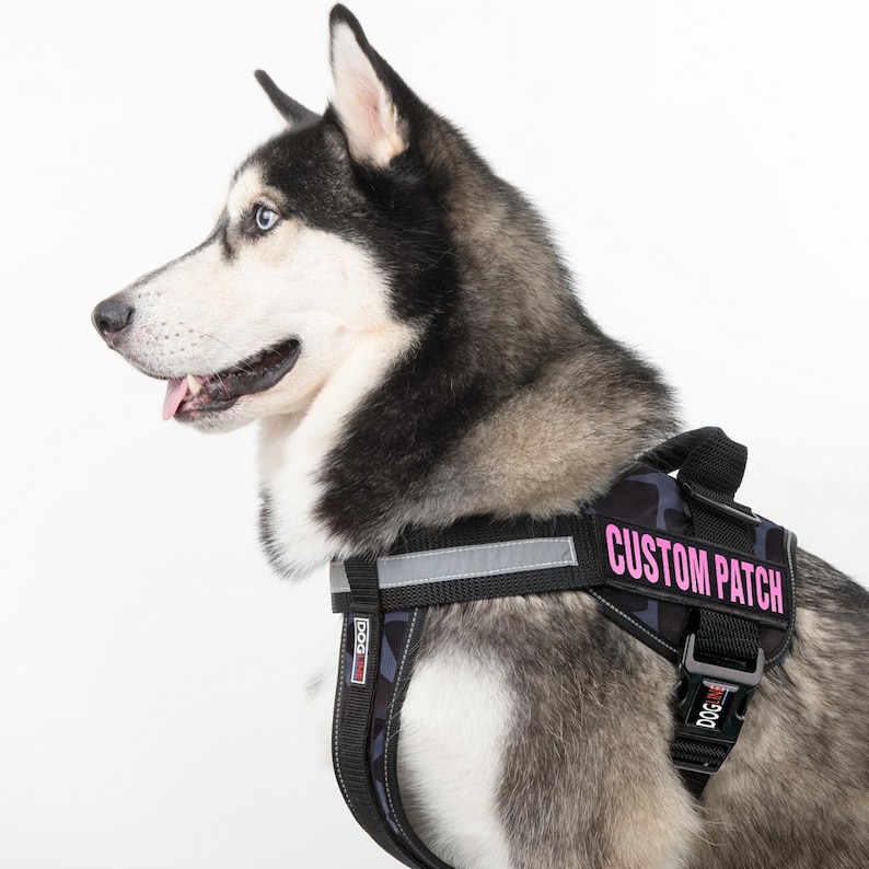 以下が含まれることがあります： 黒とグレーの迷彩柄の犬用ハーネスで、ピンクの「CUSTOM PATCH」ラベルが付いています。ハーネスには反射ストリップと、「DOG」と書かれた黒いバックルが付いています