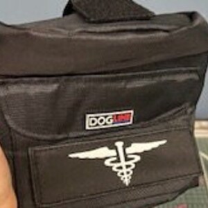 May include: Black nylon fanny pack with a rectangular front pocket and a smaller top pocket. The pack features a white medical symbol and the text "DOG LINE" in a red and blue logo.