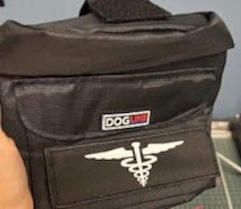 May include: Black nylon fanny pack with a rectangular front pocket and a smaller top pocket. The pack features a white medical symbol and the text "DOG LINE" in a red and blue logo.