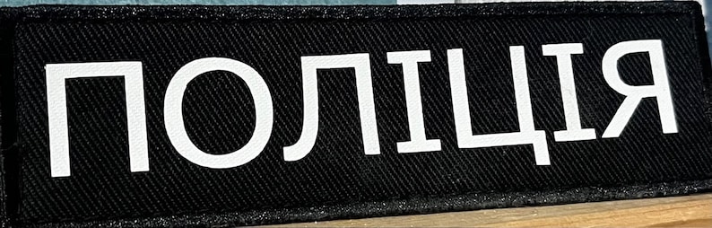 Może przedstawiać: Czarny naszywka z białym tekstem, kt&oacute;ry brzmi "ПОЛИЦИЯ", co jest rosyjskim słowem oznaczającym "Policja".