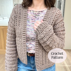 May include: A light brown open-front crochet cardigan with long sleeves and a textured stitch pattern. The garment is worn over a multicolored speckled knit top and blue jeans with a brown leather belt. A circular label reads Crochet Pattern.