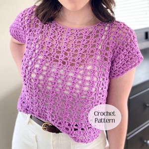 May include: A short-sleeved, purple crochet top featuring an open, airy stitch pattern with a round neckline. The garment is layered over a white base layer and includes a circular white graphic overlay with the text Crochet Pattern.