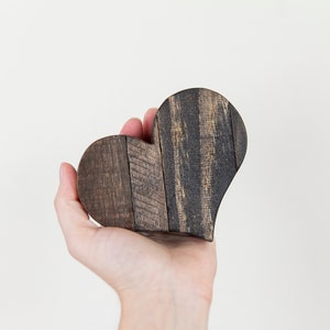 May include: A hand holds a dark brown wooden heart-shaped ornament. The heart is made of wood planks that have been stained a dark brown color.