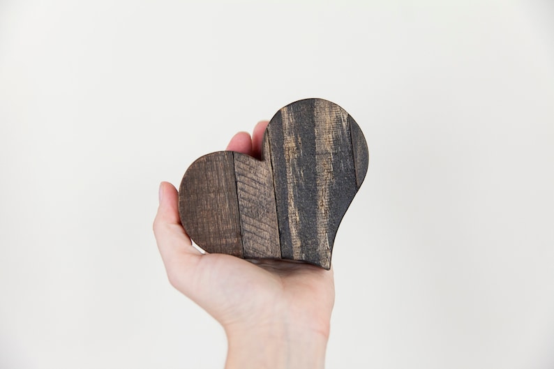 May include: A hand holds a dark brown wooden heart-shaped ornament. The heart is made of wood planks that have been stained a dark brown color.