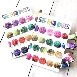 Könnte beinhalten: Set mit bunten Haargummis mit weißem Hintergrund und dem Text "She Who Dares" und "Design". Die Haargummis haben ein sich wiederholendes Muster aus schillernden Würfeln.