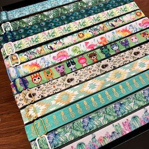 May include: A collection of colorful fabric wristbands with various patterns, including tropical leaves, pineapples, and animal characters. The wristbands have gold metal clasps.