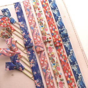 May include: A set of six colourful fabric bookmarks with gold metal clips. The bookmarks feature various Japanese-inspired patterns, including floral designs, waves, and geometric shapes. The bookmarks are perfect for adding a touch of elegance to any book.
