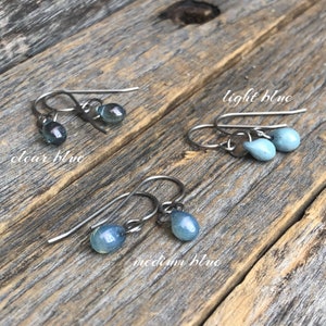 May include: Four pairs of earrings with teardrop-shaped glass beads in shades of blue. The earrings are made with silver-toned hooks. The colors are labeled "clear blue", "light blue", "medium blue", and "dark blue".